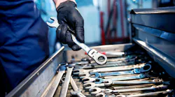 a person in mechanic's coveralls holds a ratcheting wrench above an open tool drawer of wrenches
